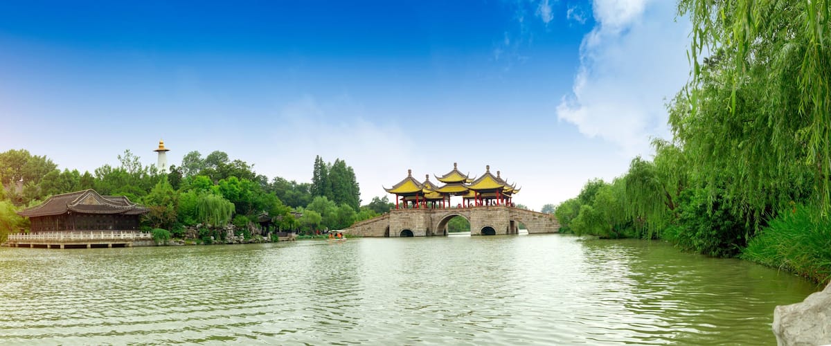 A traditional Chinese pavilion with yellow roofs by a calm river, surrounded by lush greenery and a clear blue sky.