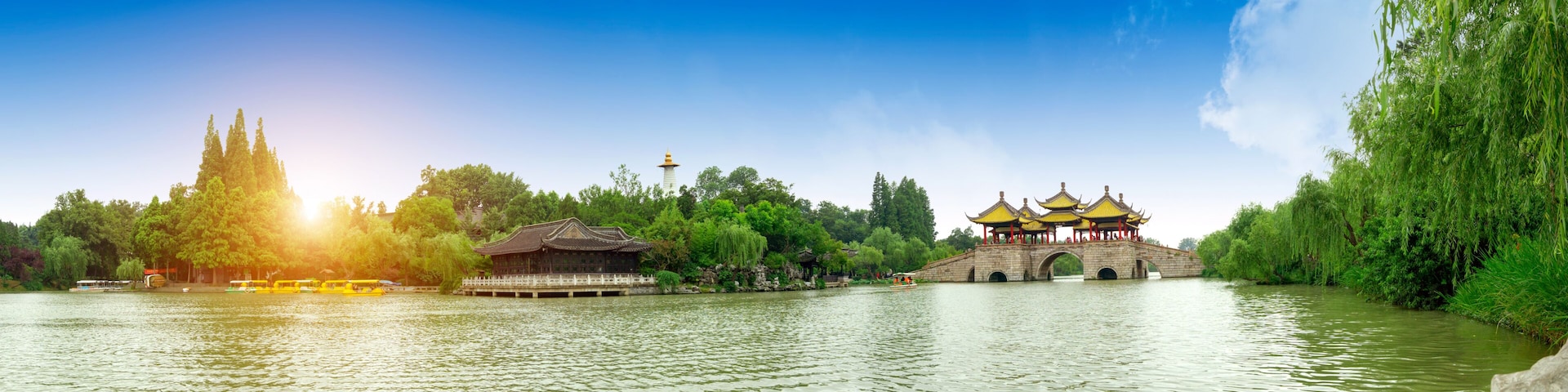 A traditional Chinese pavilion with yellow roofs by a calm river, surrounded by lush greenery and a clear blue sky.