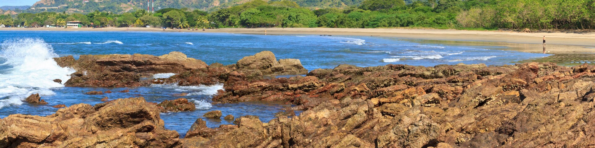 Litoral rochoso com ondas, uma praia de areia e colinas verdes ao fundo.