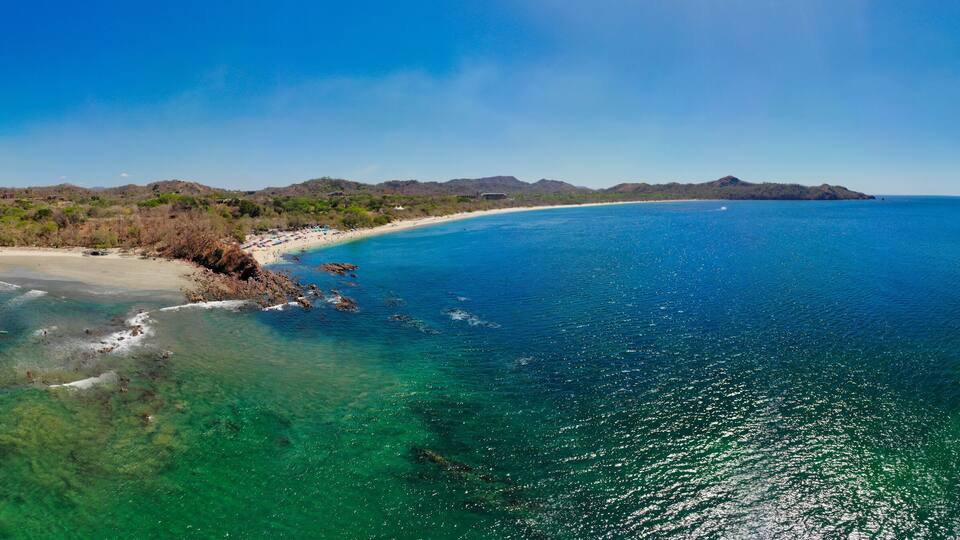A coastal landscape with clear turquoise waters, a sandy beach, and distant mountains.