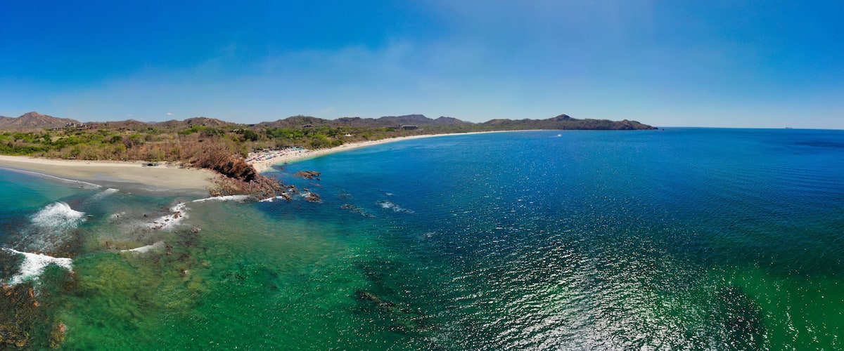 A coastal landscape with clear turquoise waters, a sandy beach, and distant mountains.
