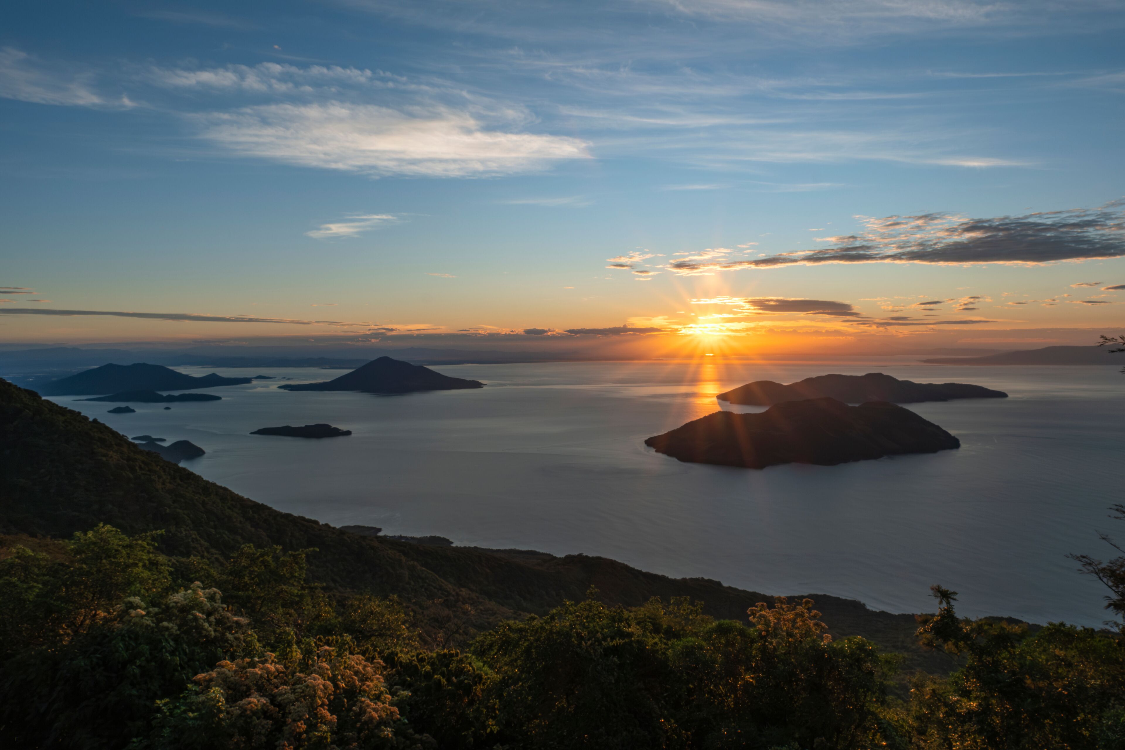 A sunset over a calm sea with several islands, surrounded by hills.