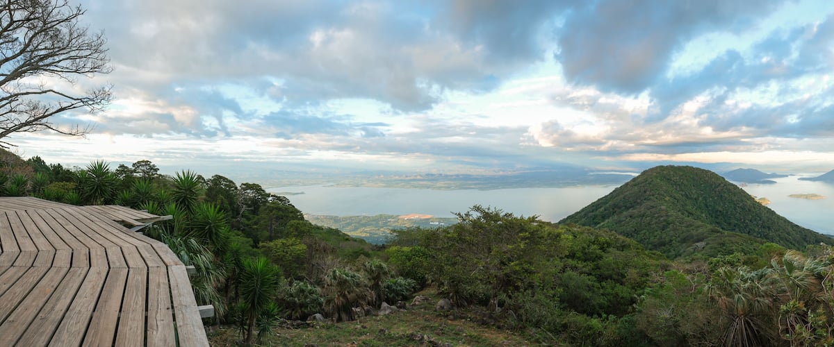 A mountainous landscape with a wooden viewing platform, lush greenery, and a distant view of a large body of water.