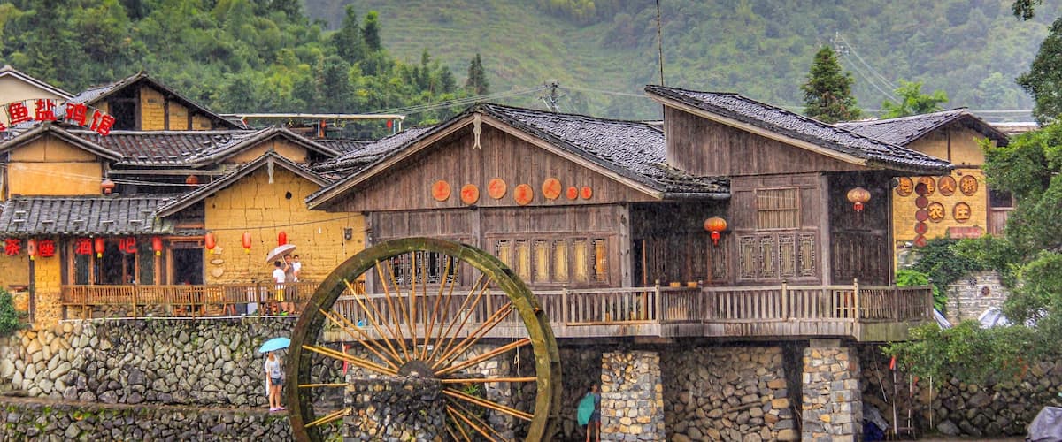 Traditional wooden buildings with large water wheels, stone walls, and lush green hills in the background.