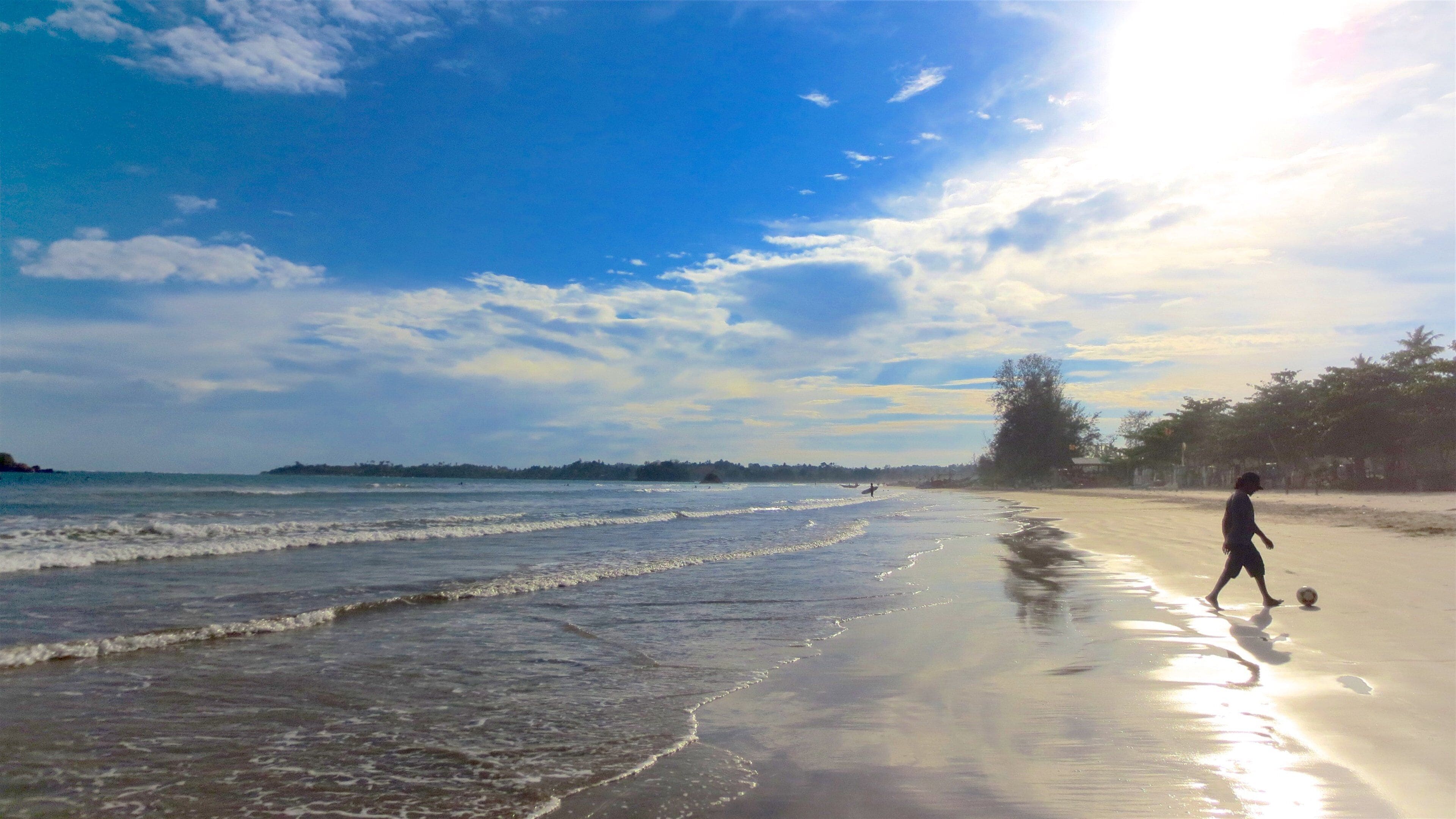 A beach with waves, a person playing soccer, and trees in the background.