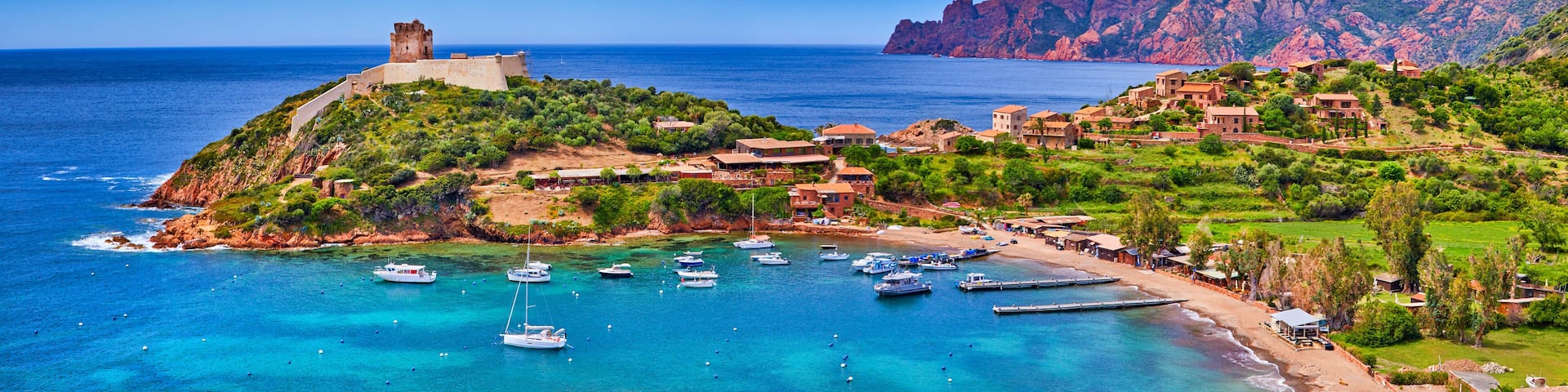 A coastal town with a historic castle, clear turquoise waters, and mountainous terrain in the background.