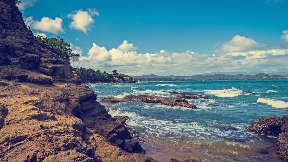 A rocky coastline with waves crashing against the shore, a clear blue sky, and distant mountains.