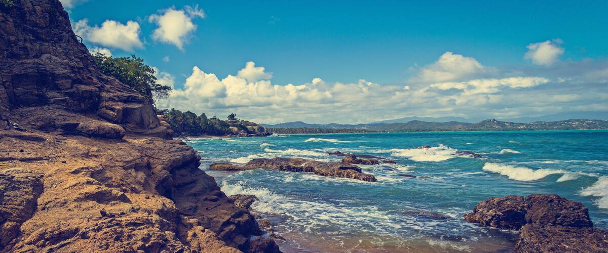 A rocky coastline with waves crashing against the shore, a clear blue sky, and distant mountains.
