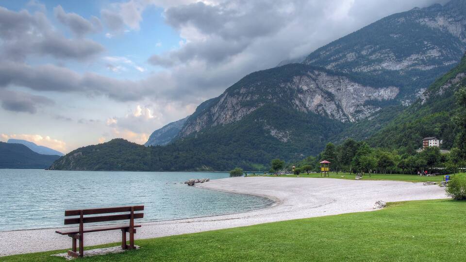 A lakeside park with a bench, grassy areas, and mountains in the background.