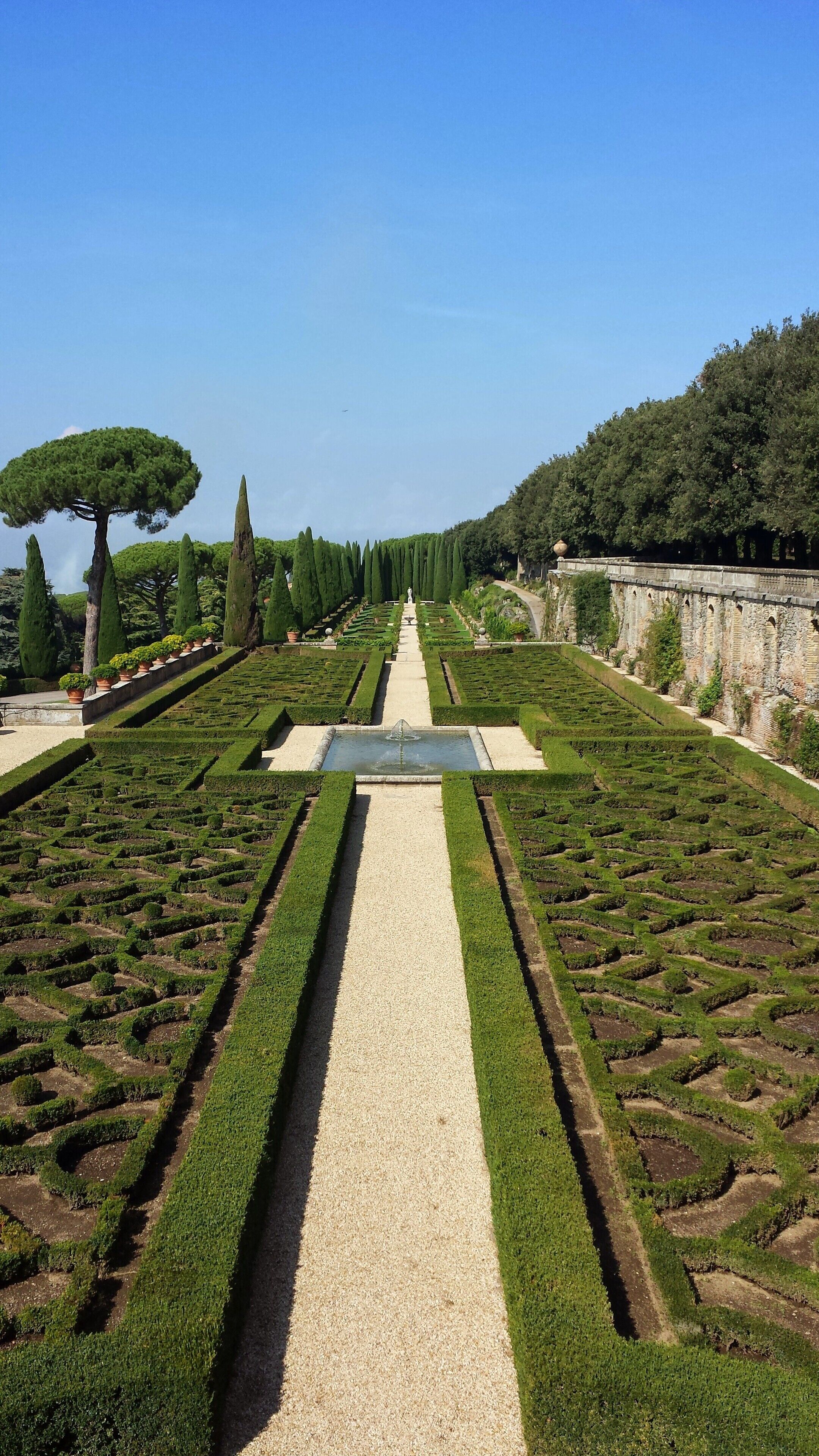 A formal garden with geometrically arranged hedges, a central gravel path, and a fountain.