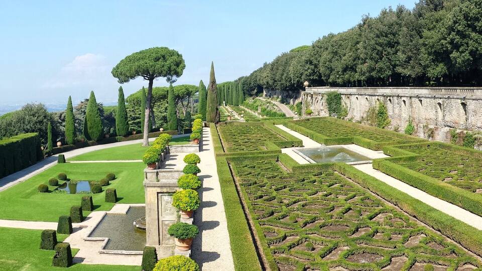 A well-manicured garden with geometric hedges, a stone bridge, and a large tree.