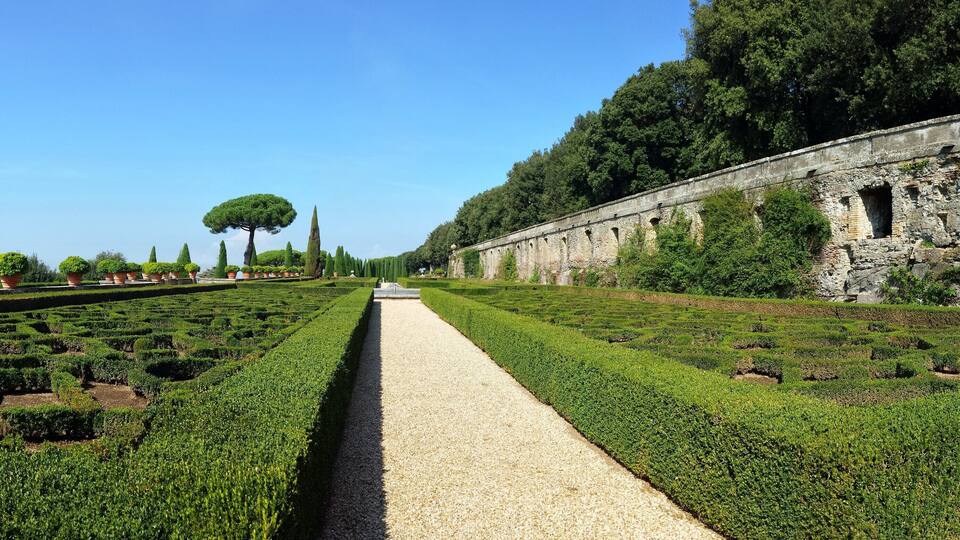 A formal garden with neatly trimmed hedges, a central gravel path, and a historic stone wall with arched windows.