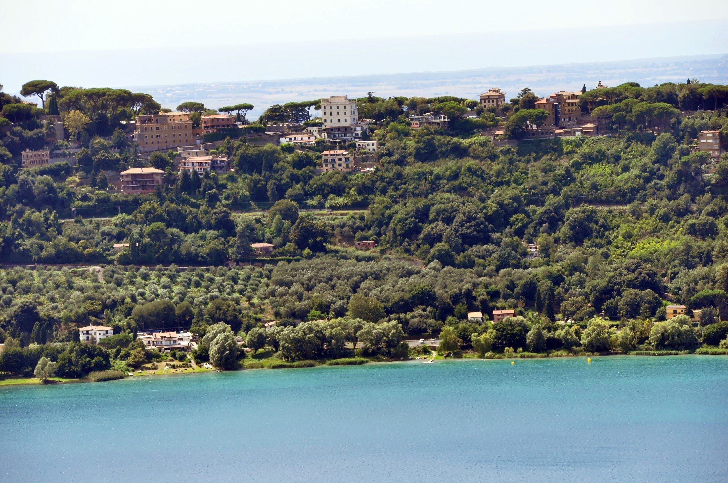 A hillside town with buildings nestled among trees and a lake in the foreground.