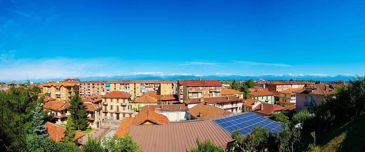 A town with red-tiled roofs, a mountain range in the background, and a clear blue sky.