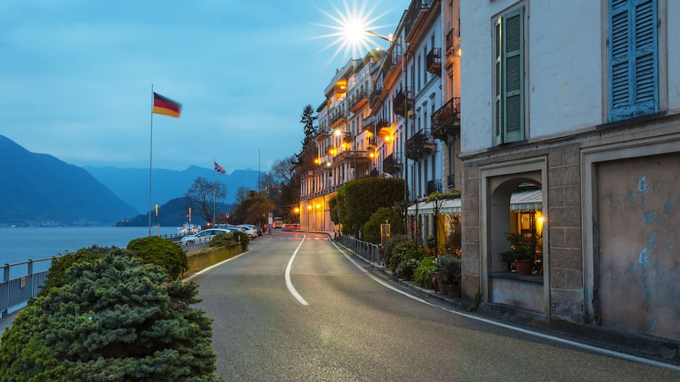 A waterfront street with buildings, a flag, and mountains in the background.