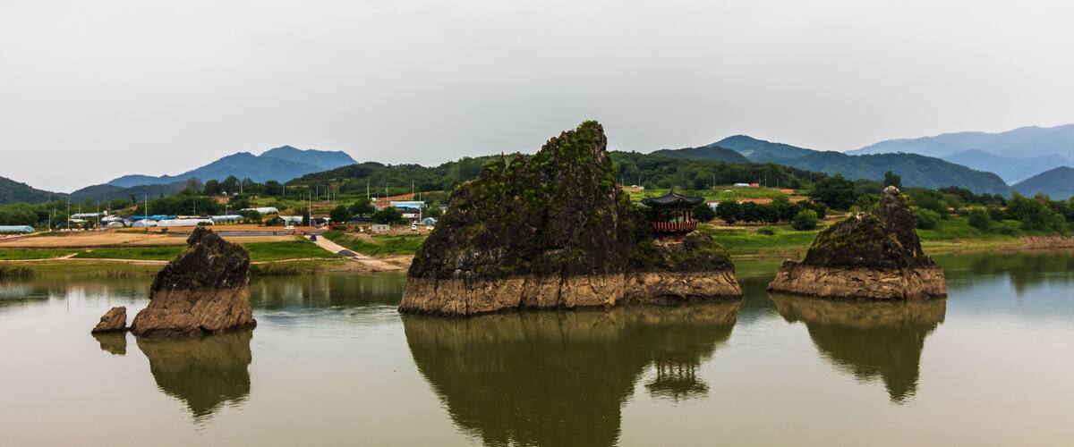 A calm river with three prominent rock formations in the middle. In the background, there are mountains and a small village with traditional houses.