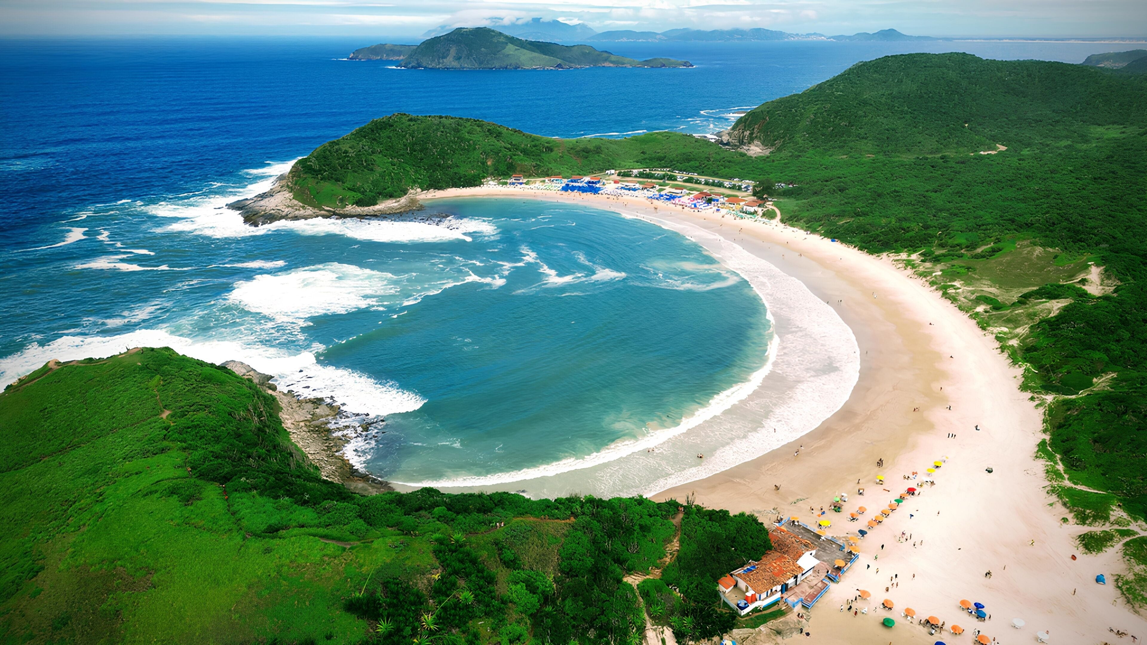 A coastal landscape with a curved sandy beach, lush green hills, and a small island in the distance.