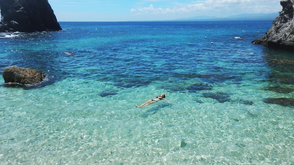 A clear blue sea with a person swimming, rocky formations, and a distant mountain range.