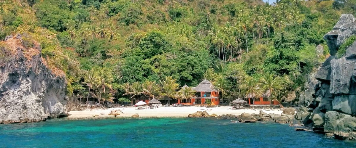 A tropical beach with clear turquoise waters, a rocky coastline, and lush green hills in the background.
