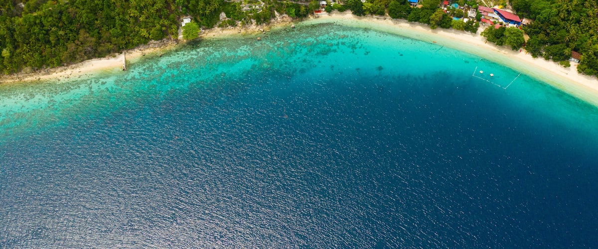 An aerial view of a turquoise beach with a dense forest and scattered buildings.
