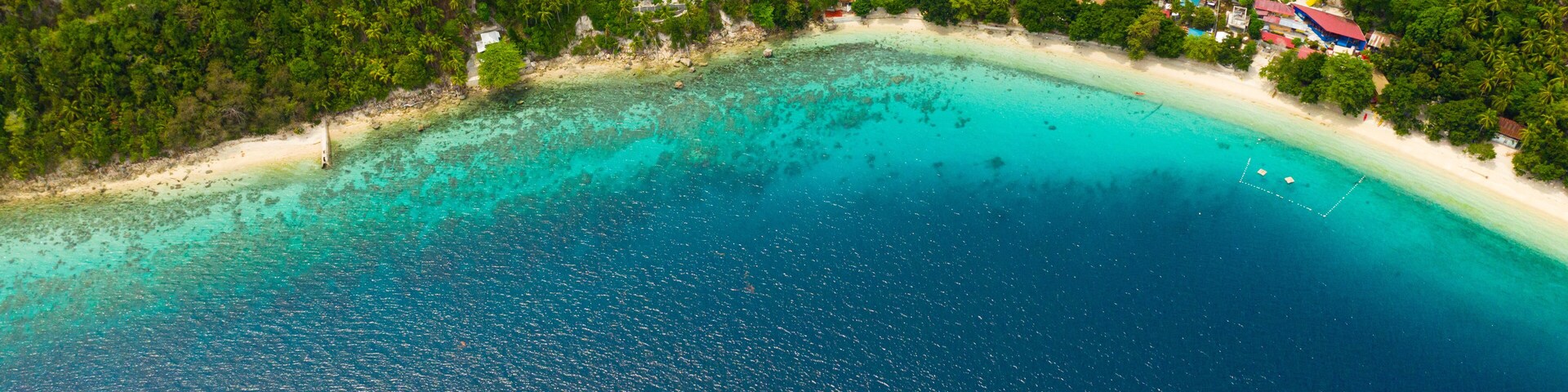 An aerial view of a turquoise beach with a dense forest and scattered buildings.