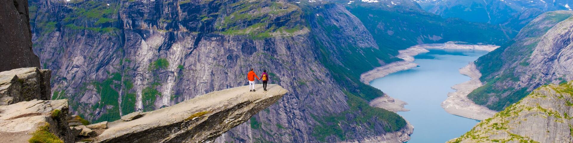Two hikers stand on a dramatic cliff edge, overlooking Trolltunga, Norway majestic fjords and lush valleys. The sky is partly cloudy, creating a perfect backdrop for this awe-inspiring landscape.