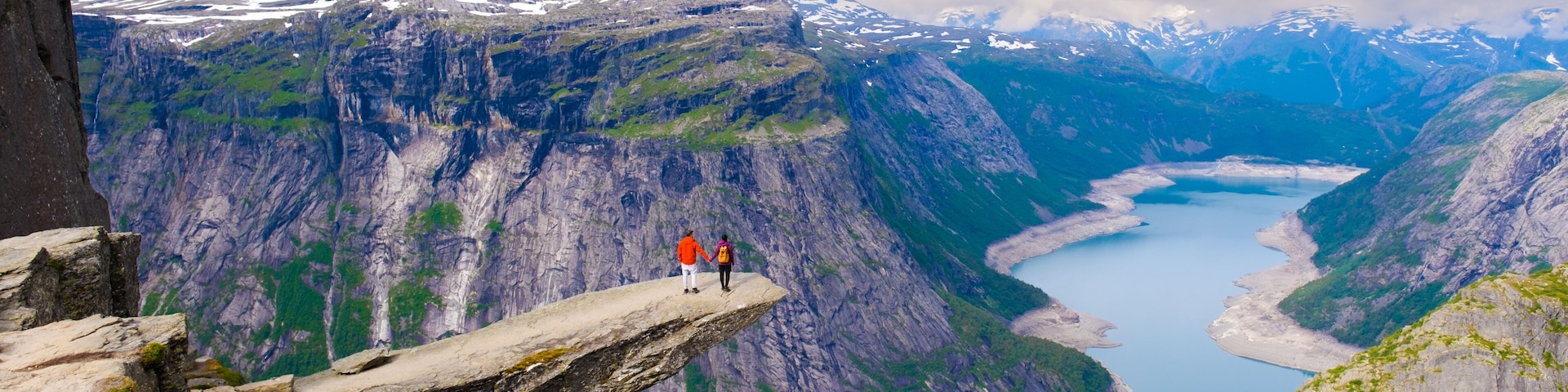 Two hikers stand on a dramatic cliff edge, overlooking Trolltunga, Norway majestic fjords and lush valleys. The sky is partly cloudy, creating a perfect backdrop for this awe-inspiring landscape.