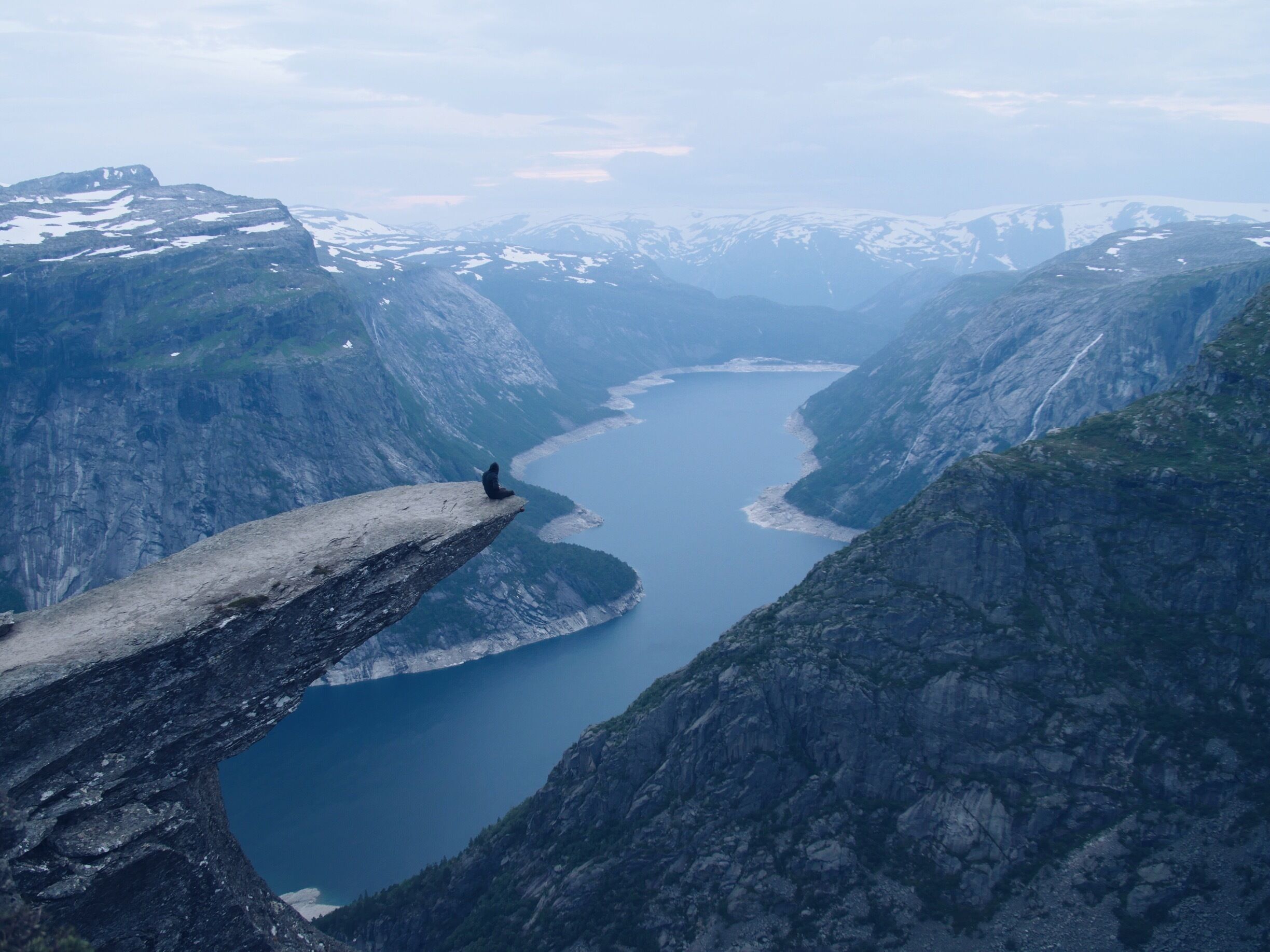 A person sitting on a narrow ledge overlooking a deep valley with a lake.