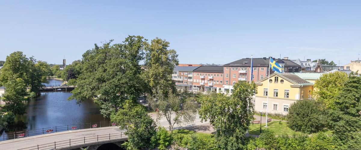 A river with a bridge, trees, and buildings including a yellow house with a Swedish flag.