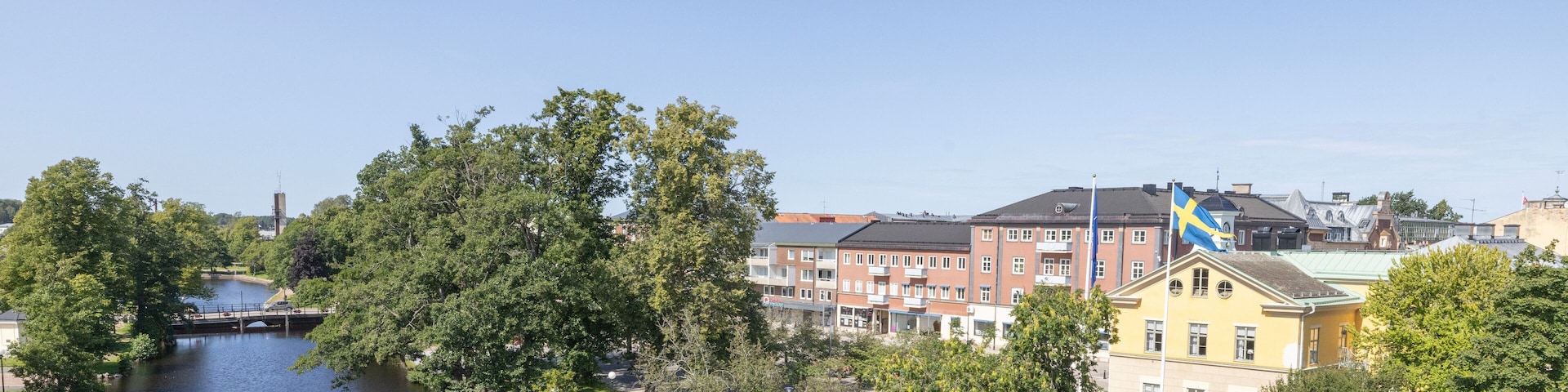 A river with a bridge, trees, and buildings including a yellow house with a Swedish flag.