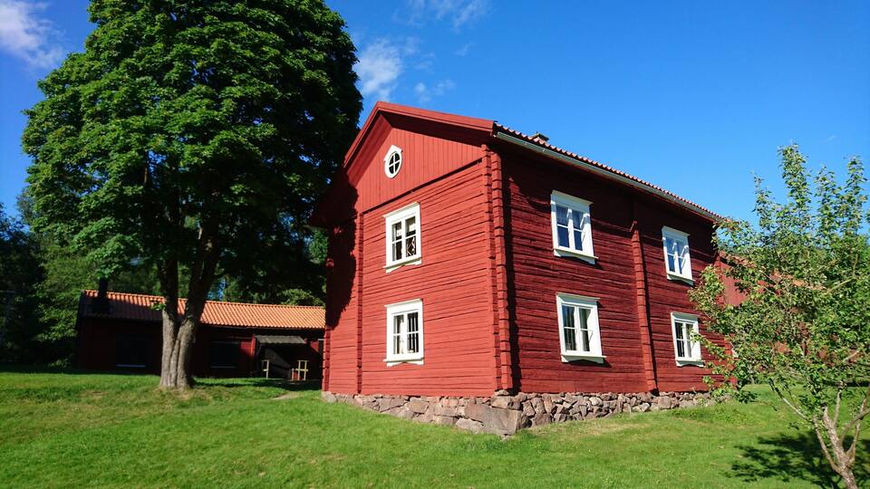A red wooden house with a stone foundation, surrounded by green grass and trees.