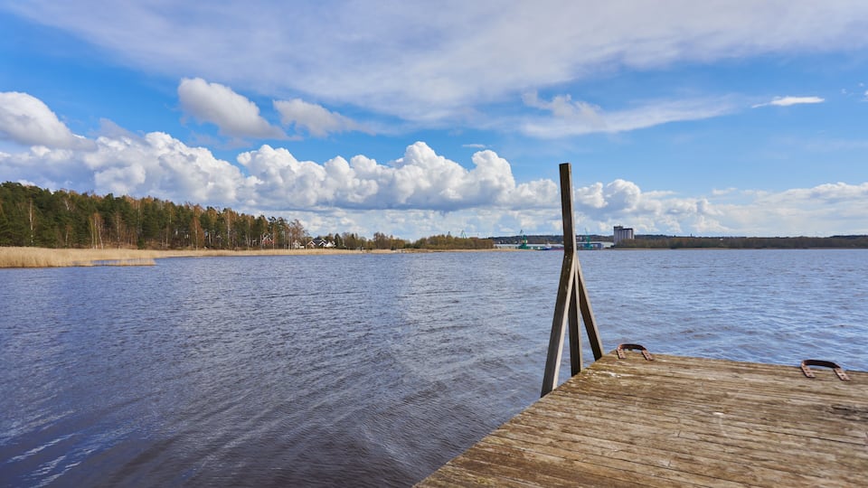 A wooden dock extending into a calm lake with a forested shoreline and industrial structures in the distance.