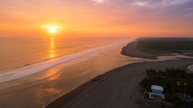 Un coucher de soleil sur une plage, avec des vagues, une côte lointaine et quelques bâtiments.