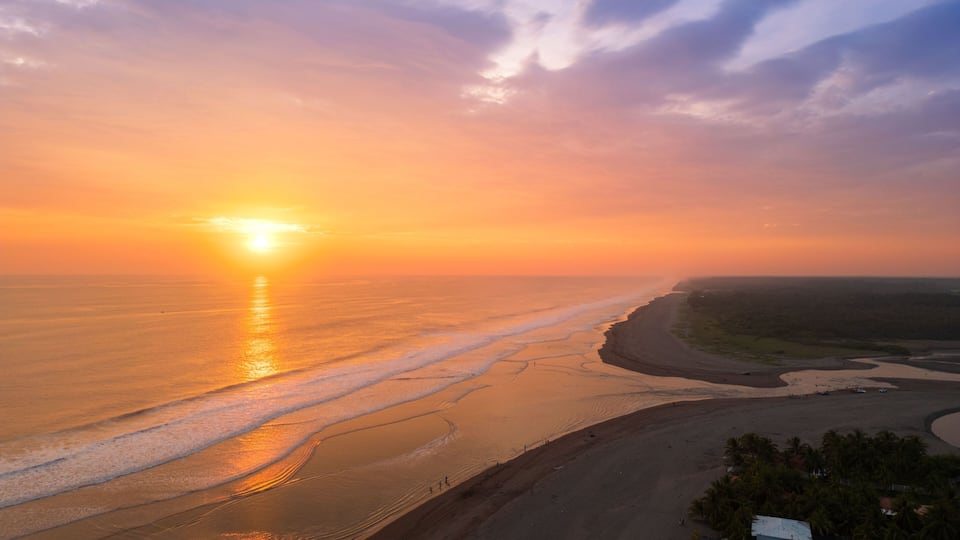 A sunset over a beach with waves, a distant shoreline, and a few buildings.