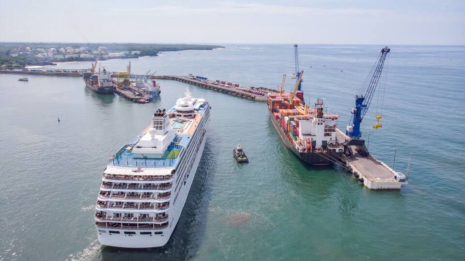 A large cruise ship docked at a port with a cargo ship loaded with containers, a tugboat, and a crane in the background.