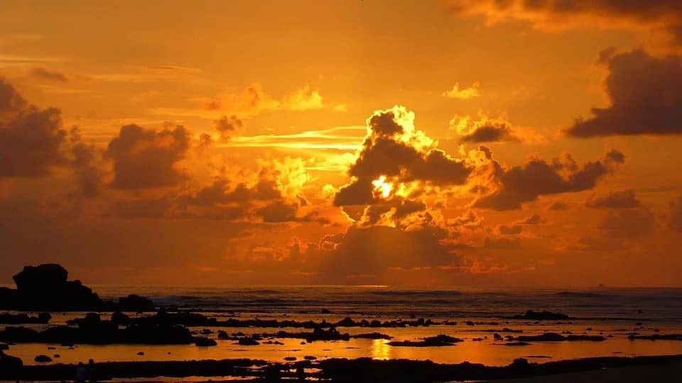A sunset over a rocky shoreline with scattered rocks and calm water reflecting the sky.