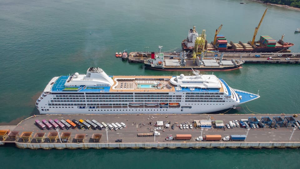 A large cruise ship docked at a port with a cargo ship nearby, a pier with parked vehicles, and a distant shoreline with buildings.