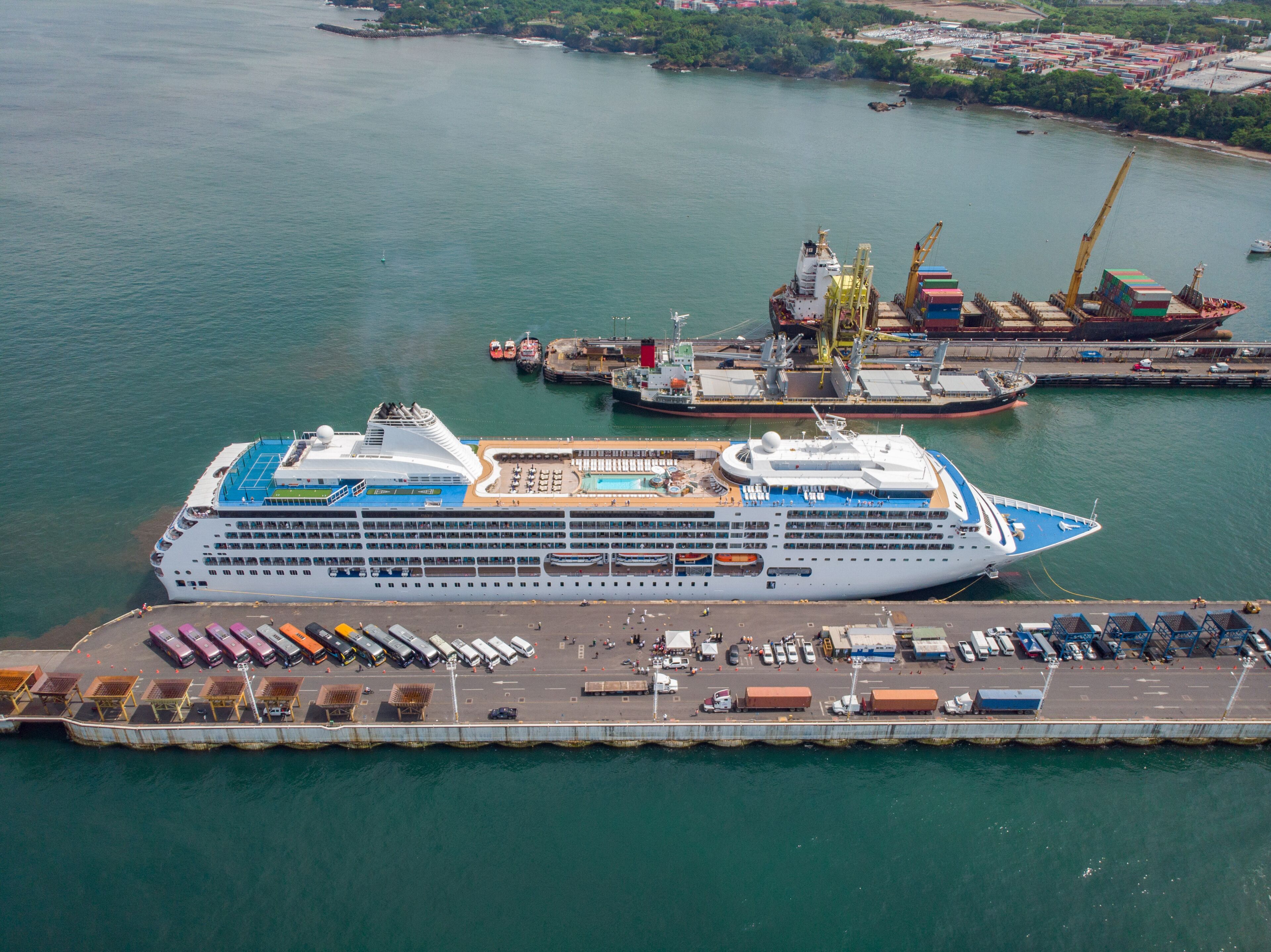 A large cruise ship docked at a port with a cargo ship nearby, a pier with parked vehicles, and a distant shoreline with buildings.