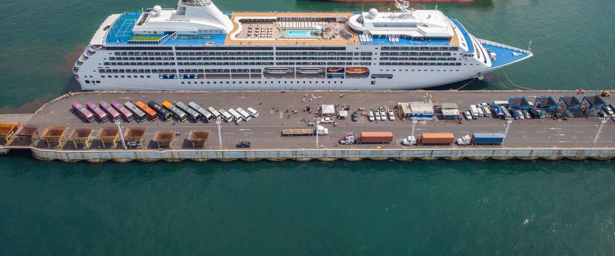 A large cruise ship docked at a port with a cargo ship nearby, a pier with parked vehicles, and a distant shoreline with buildings.