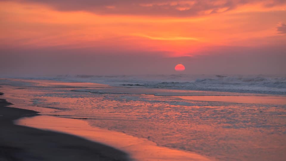 A sunset over a beach with waves and a calm sea.