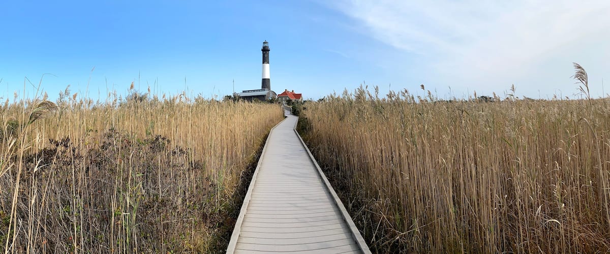 A wooden boardwalk leading to a lighthouse in a coastal area with tall grasses.