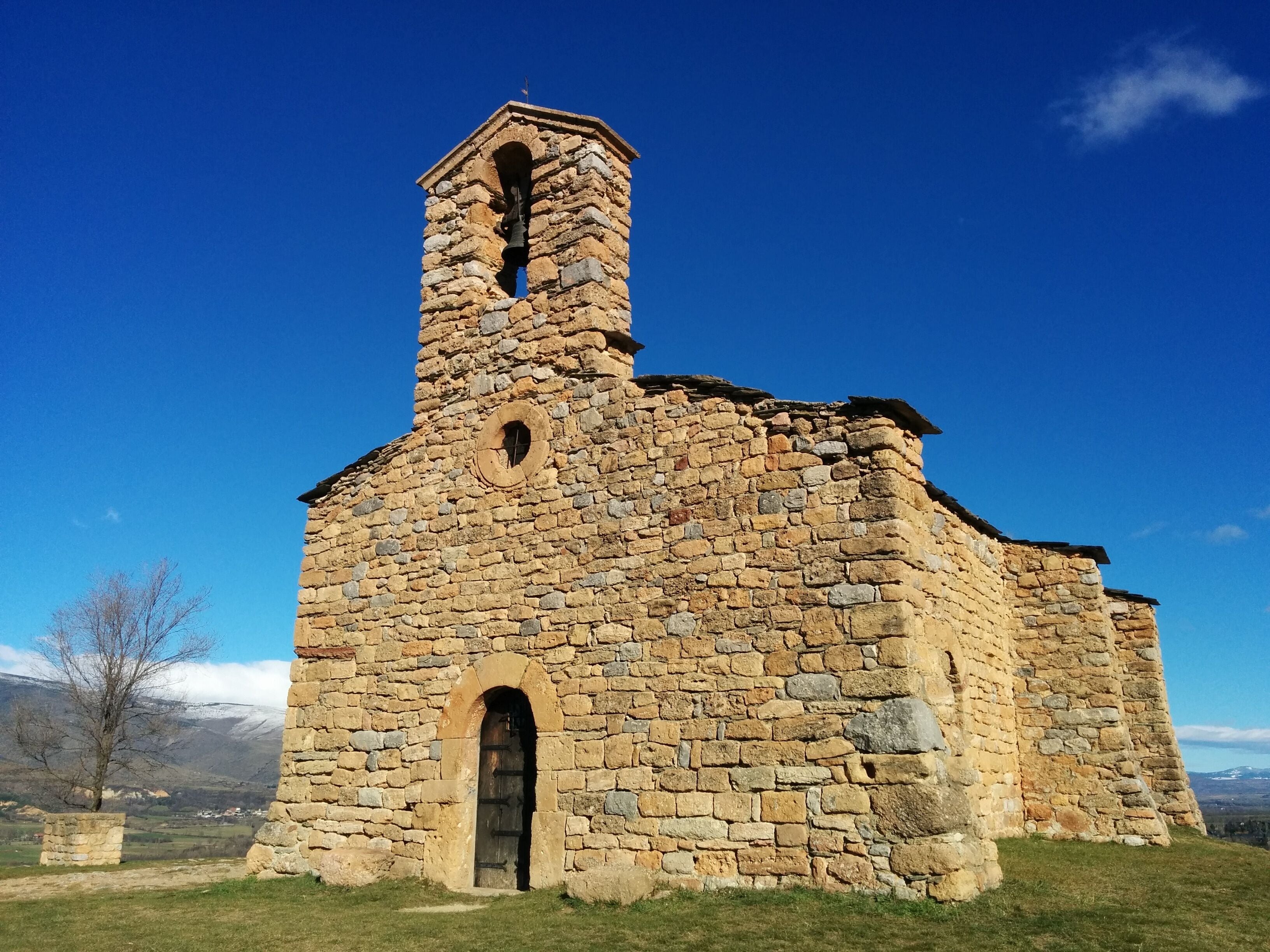 A stone church with a bell tower and arched door, set against a clear blue sky.