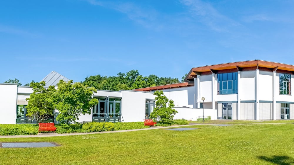 Une maison moderne à deux étages, avec un toit rouge, entourée d’une pelouse bien entretenue et d’arbres.