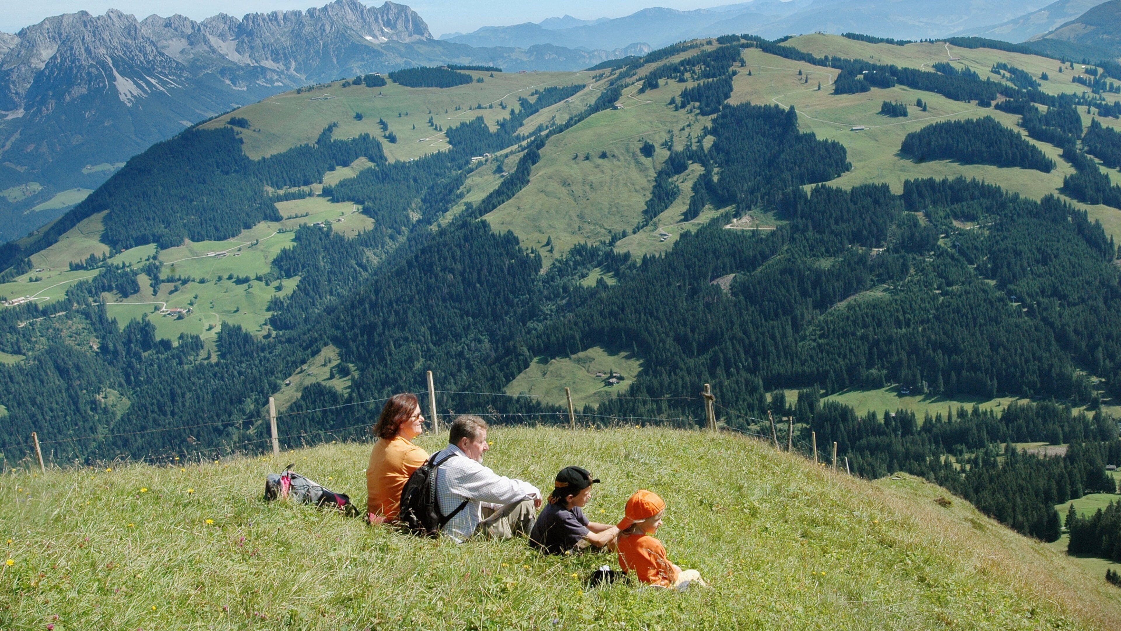 Una familia sentada en una colina cubierta de hierba, con un paisaje montañoso al fondo.