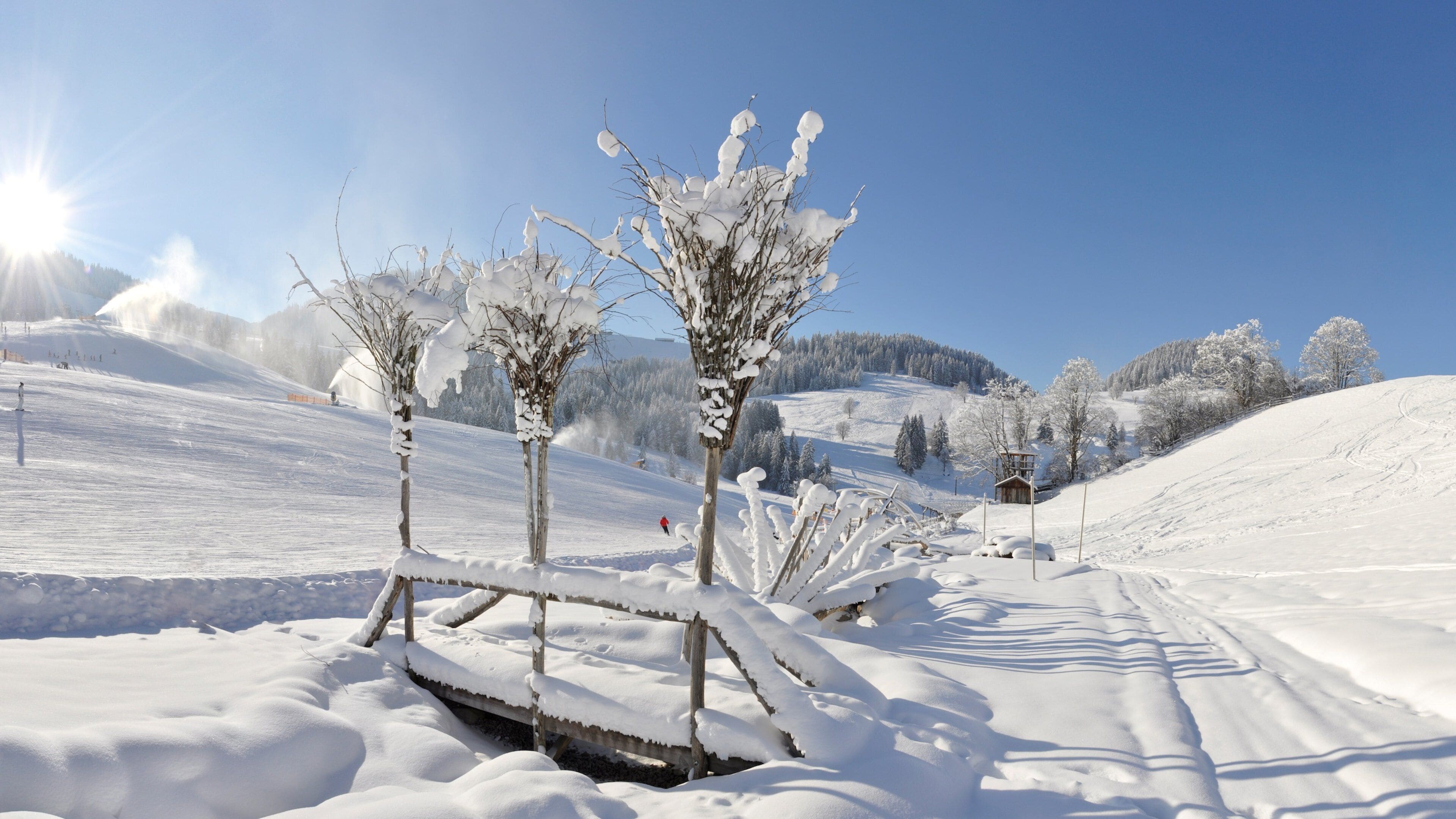 A snow-covered landscape with a wooden bridge, trees, and a ski slope.