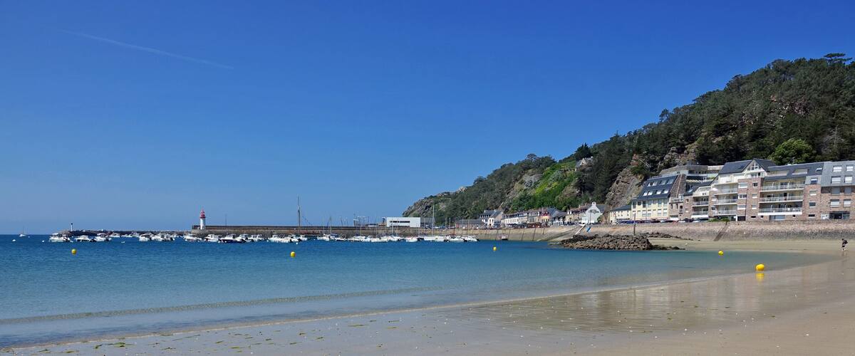 A coastal town with a clear blue sky, a sandy beach, a rocky shoreline, and a lighthouse in the distance.