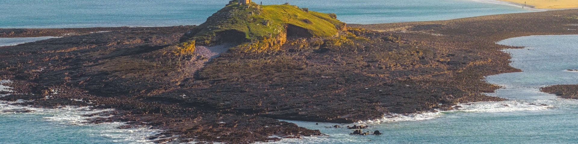 A coastal landscape with a prominent rocky island featuring a small structure, surrounded by calm waters and distant residential areas.