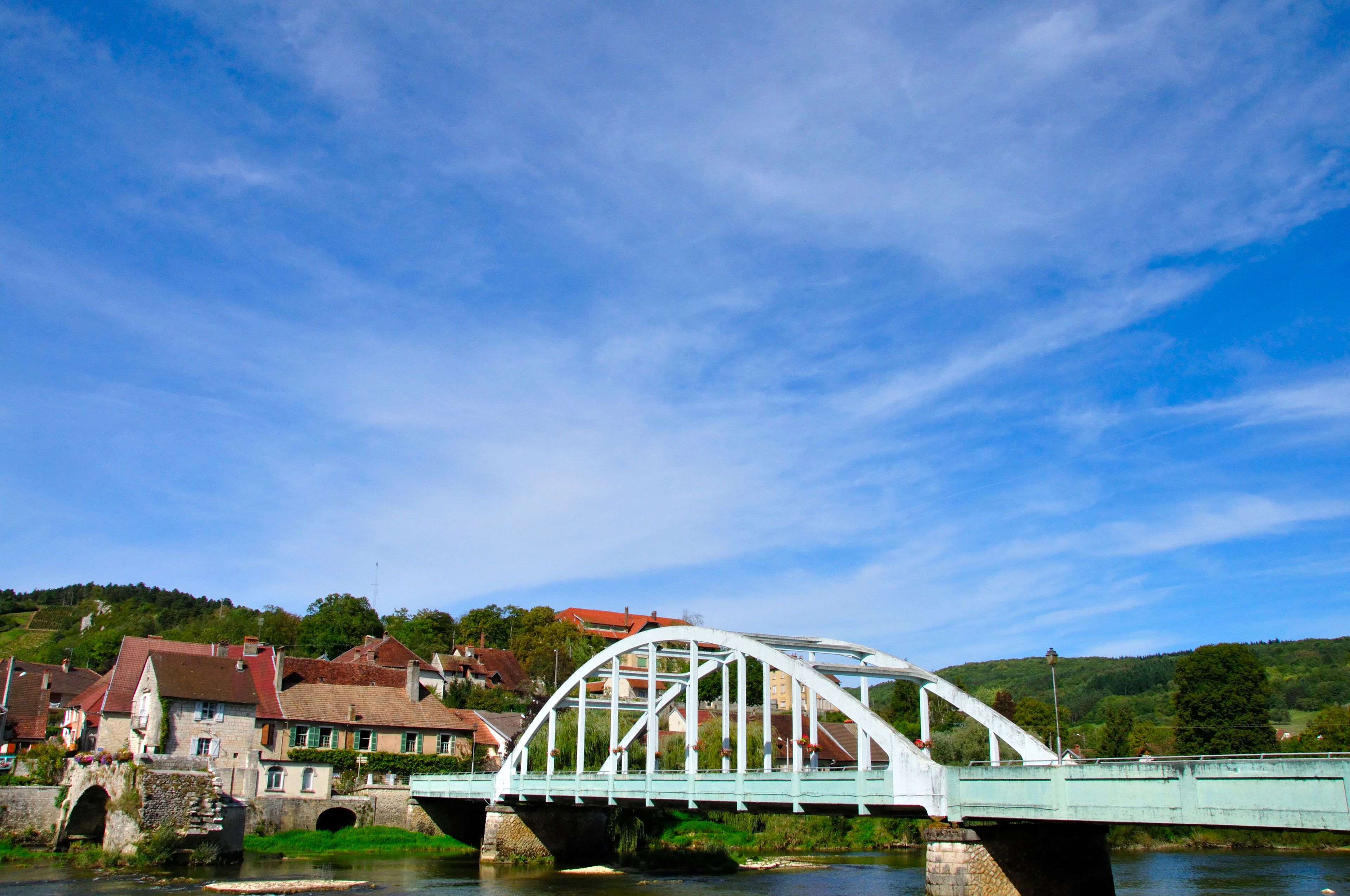 A white metal bridge over a river with houses and greenery in the background.