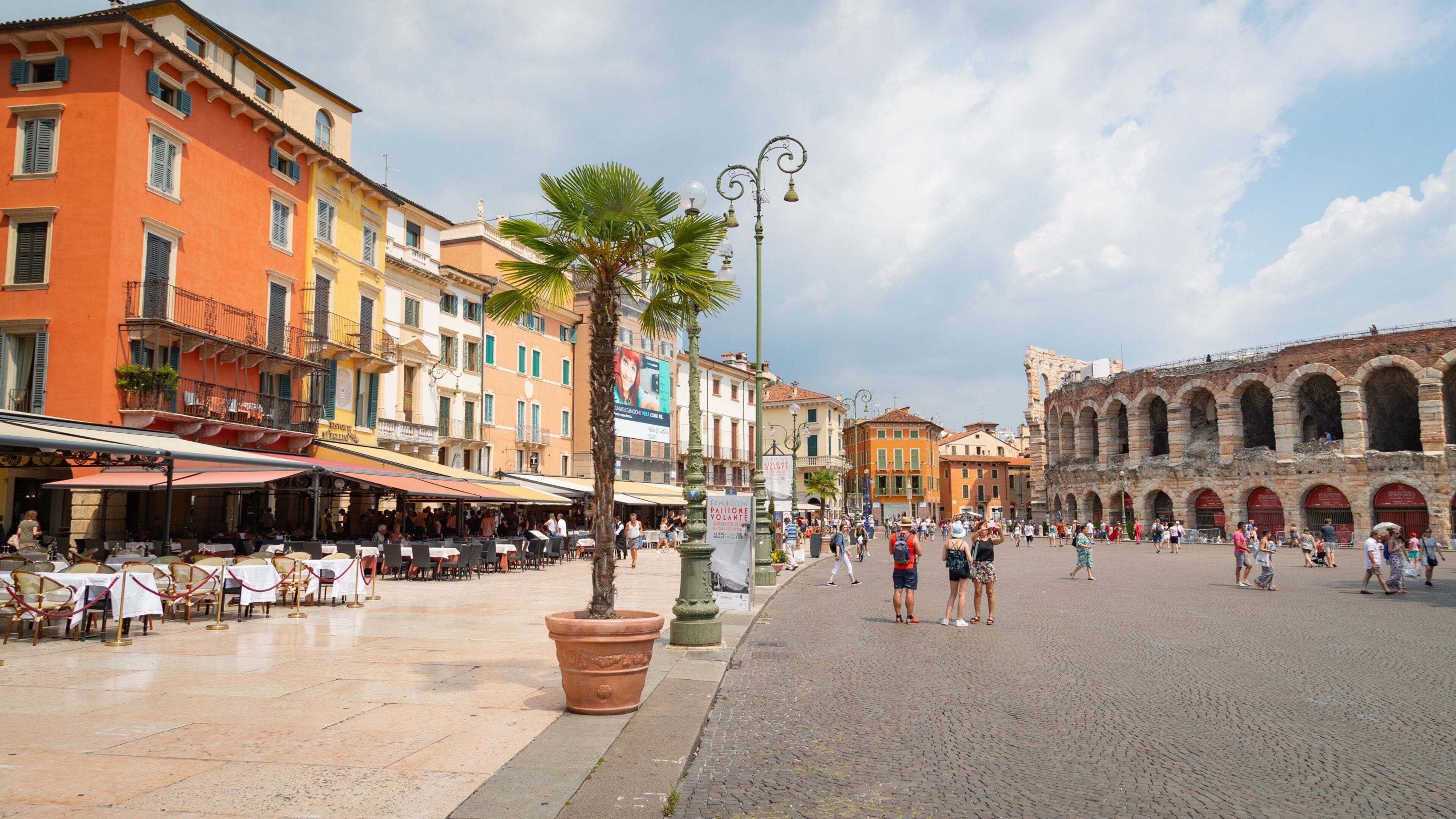 A bustling piazza with historic buildings, palm trees, and outdoor seating areas.