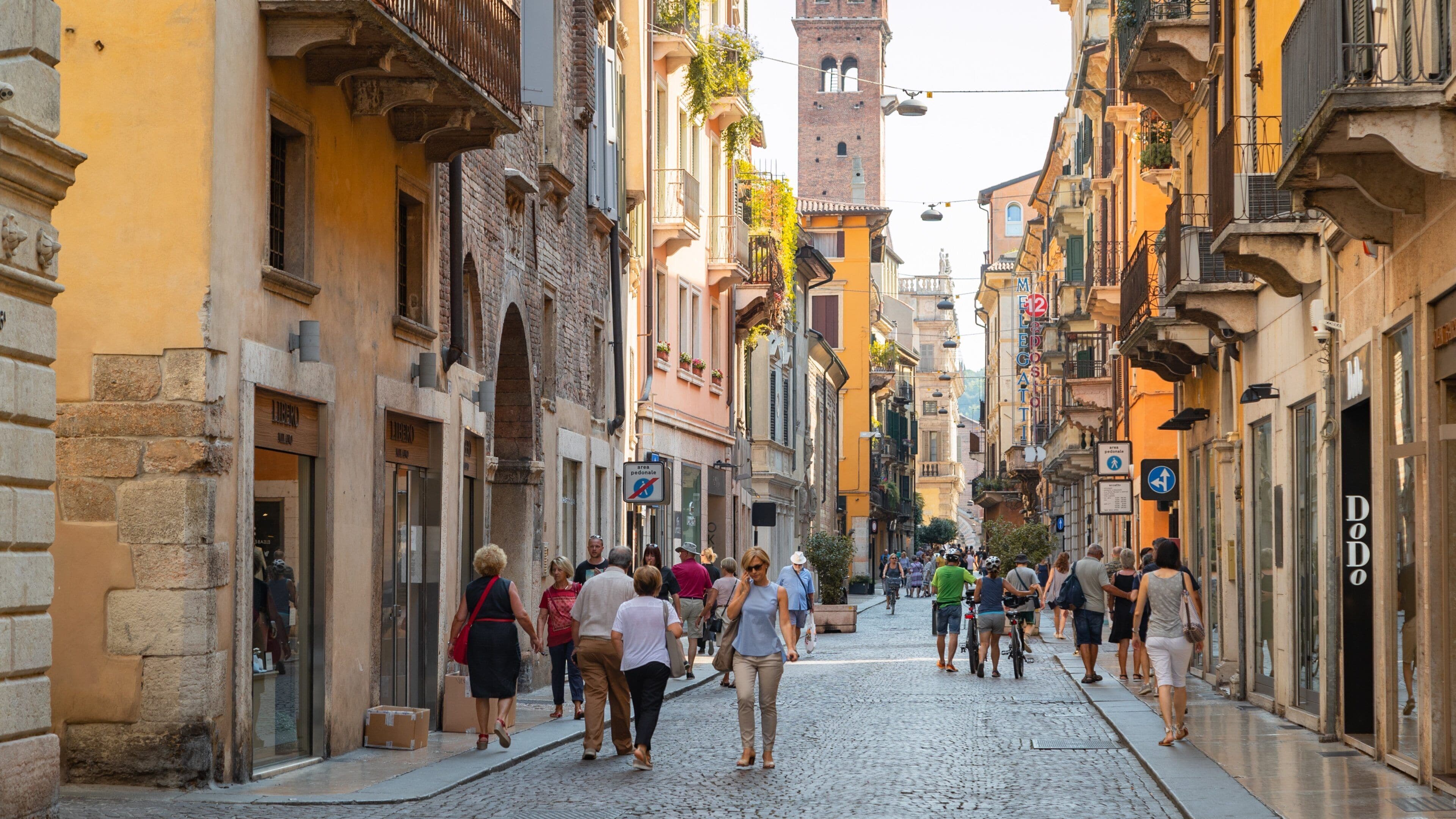A cobblestone street lined with historic buildings, a prominent clock tower, and pedestrians.